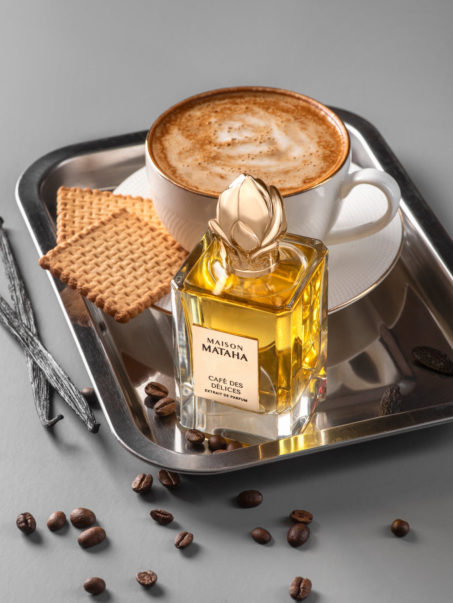 A cup of cappuccino and two rectangular biscuits rest on a silver tray beside Maison Mataha’s Café des Délices Extrait de Parfum, surrounded by coffee beans and vanilla pods on a grey surface, evoking rich coffee notes.