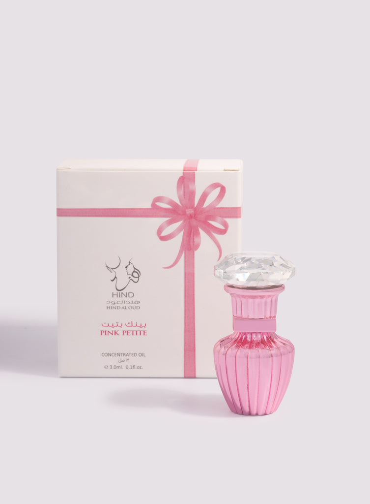 A pink perfume bottle with a faceted clear cap stands before a white box featuring a pink ribbon illustration. Jasmine and cashmere wood notes drift in the air as “Pink Petite Concentrated Oil 3ml, Hind Al Oud” is displayed in English and Arabic.
