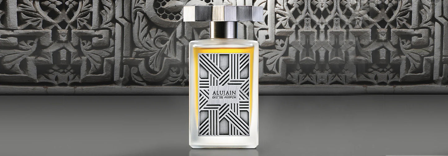 A rectangular Alujain Eau de Parfum bottle by Kajal Perfumes Paris stands on a gray surface, set against an intricate geometric and floral background. The bottle has a metallic cap and a modern, patterned label.