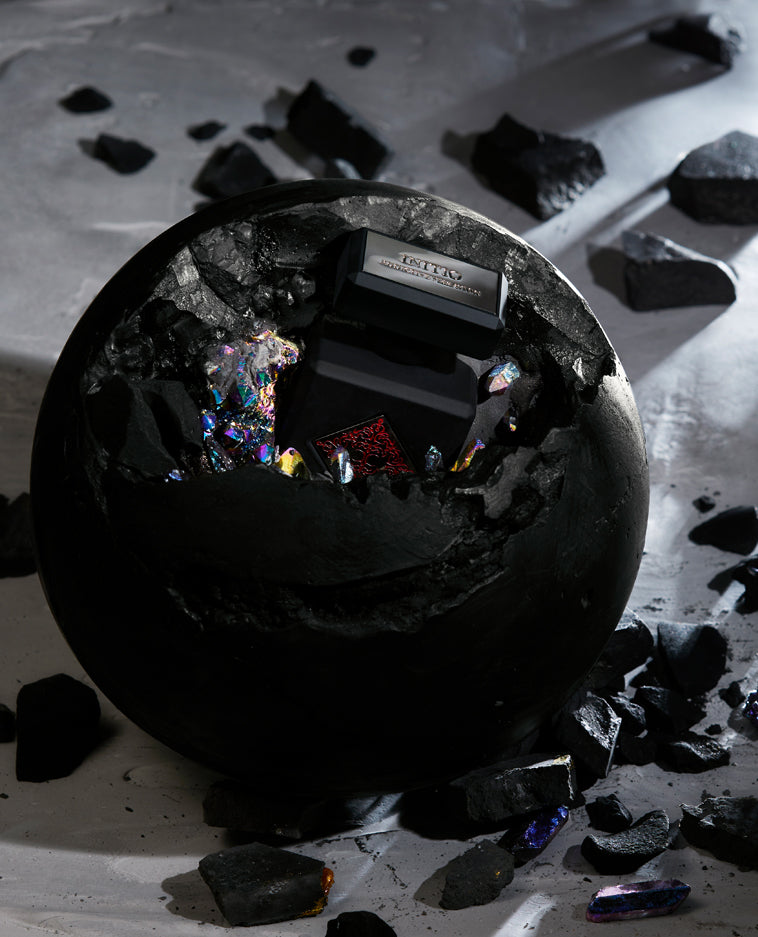 A large black sphere cracks open on a gray surface, unveiling INITIO’s Addictive Vibration Eau de Parfum boxed inside, surrounded by colorful shards as dramatic light shines and hints of musk and orange blossom linger in the air.