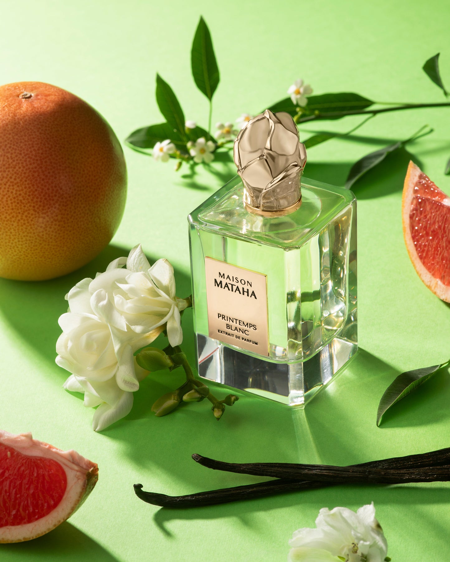 A Maison Mataha Printemps Blanc Extrait de Parfum bottle rests on a green surface, surrounded by white flowers, grapefruit halves, vanilla beans, and green leaves—evoking the fragrance’s floral notes with jasmine sambac hints.