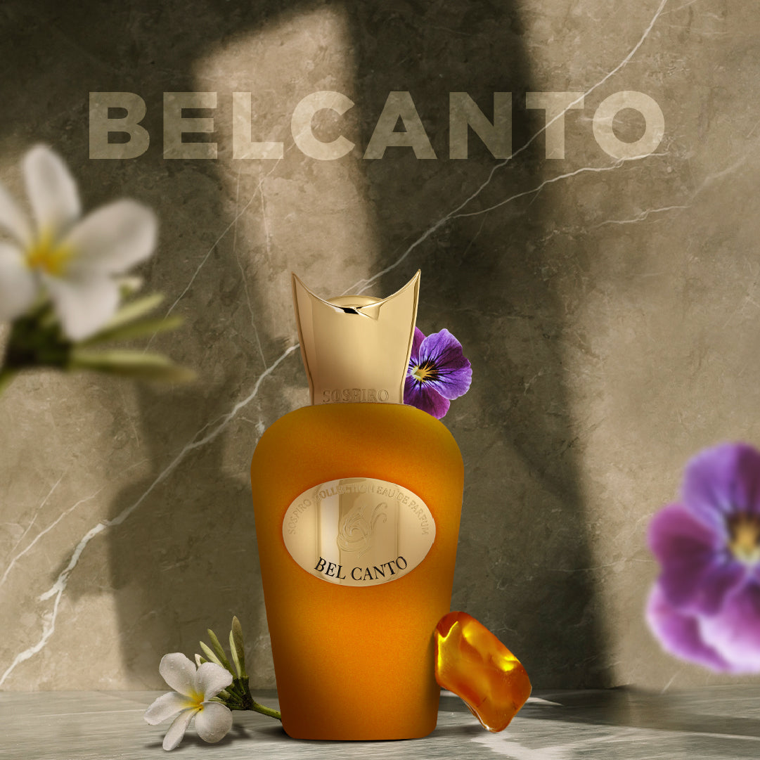 A golden bottle of Bel Canto Eau de Parfum by SOSPIRO rests on marble amidst vibrant flowers and a golden petal, evoking its floral scent with sandalwood notes. BEL CANTO is boldly displayed on the wall behind.