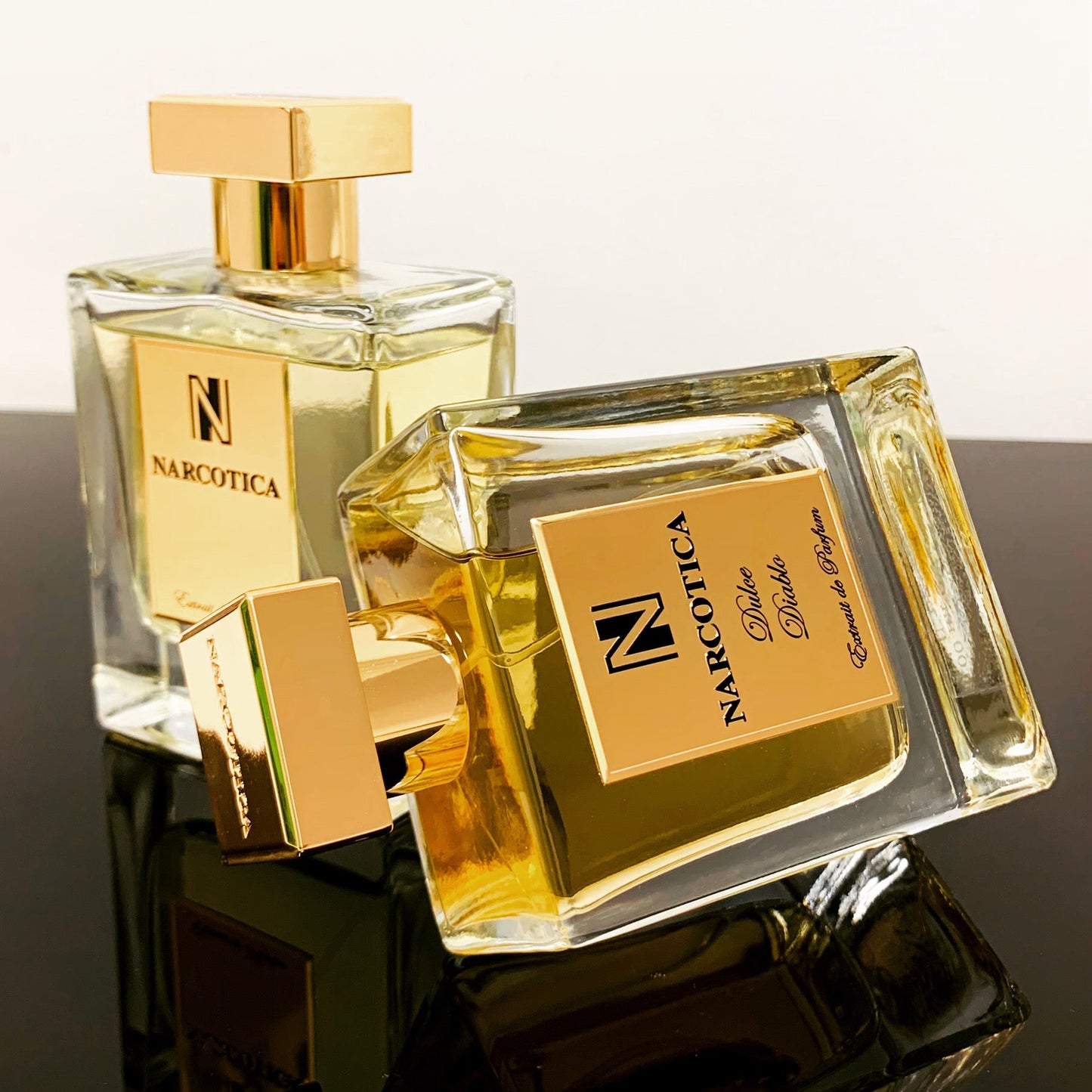 Two clear glass bottles of Dulce Diablo Eau de Parfum by Narcotica, each with gold caps and labels—one upright, one on its side—rest on a reflective black surface, evoking the decadent blend of rum perfume and Madagascar vanilla.