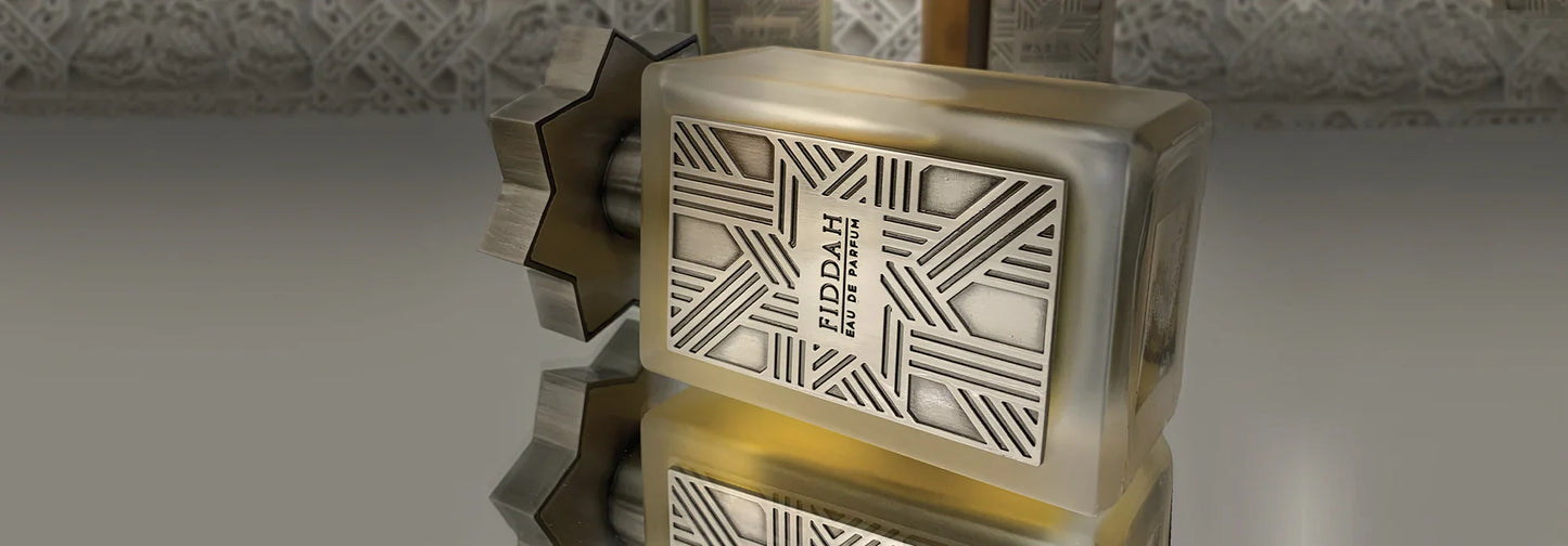 The Fiddah Eau de Parfum by Kajal Perfumes Paris rests on its side, showcasing an elegant geometric design on its silver label and a star-shaped cap, with the patterned background mirrored on its shiny surface.