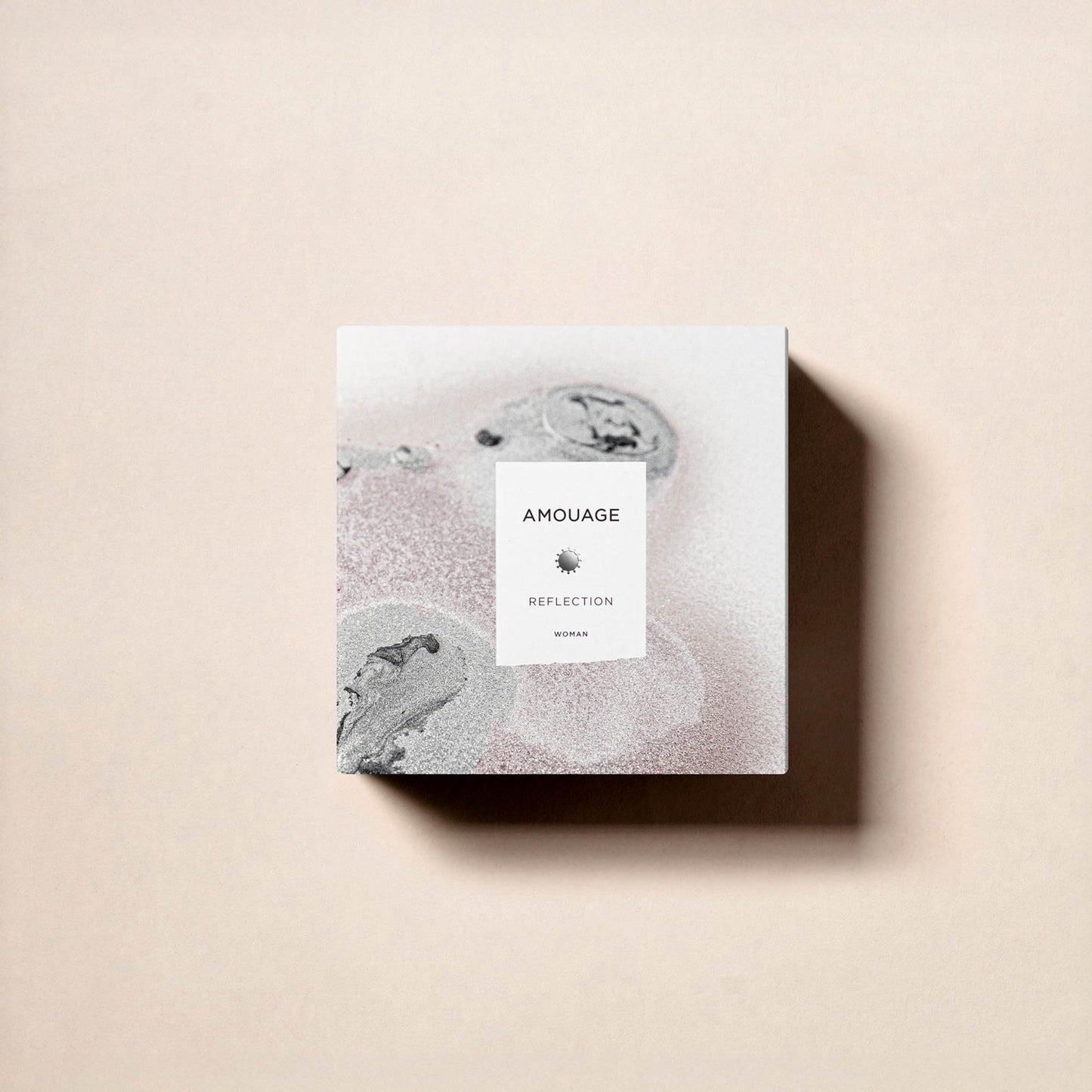 A square perfume box from Fragrapedia Haus, labeled Reflection Woman Eau de Parfum - Amouage, rests on a light beige surface. Its minimalist design features abstract gray and pale pink patterns, evoking the white floral scent inside.