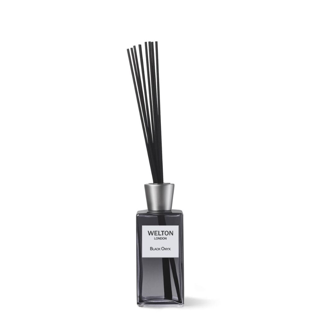 A rectangular glass bottle labeled Welton London holds the BLACK ONYX HOME FRAGRANCE DIFFUSER, featuring thin black reeds in a silver cap and releasing woody notes of vetiver, teak, and amber.