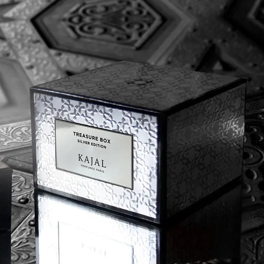A reflective silver Kajal Silver Treasure Box Discovery Kit by Kajal Perfumes Paris rests on a patterned surface, ornate designs behind it—an elegant set for luxury fragrance lovers.