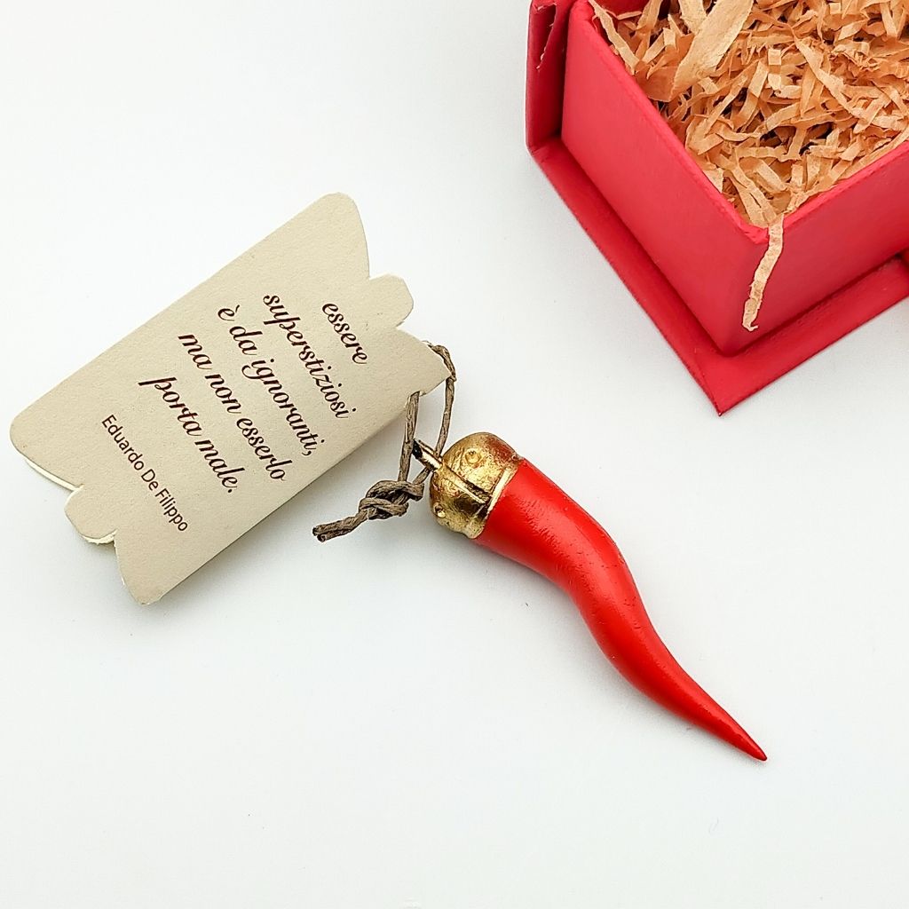 The ANTI MALOCCHIO Eau de Parfum by Nobile 1942 features a red Italian horn charm with a gold cap tied to brown string on a beige Italian-tag, beside a pink gift box with shredded paper—evoking the style of scented amulets.