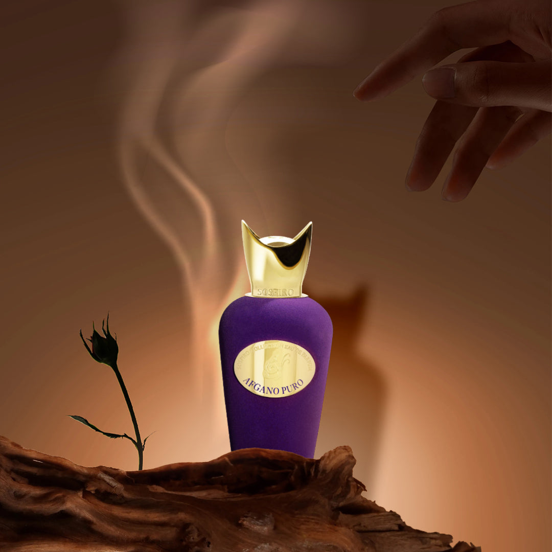 A luxurious purple and gold bottle of Afgano Puro Eau de Parfum by SOSPIRO sits on textured wood with a rosebud and swirling mist, evoking a tobacco vanilla scent. A hand enters from the upper right against a warm, glowing background.