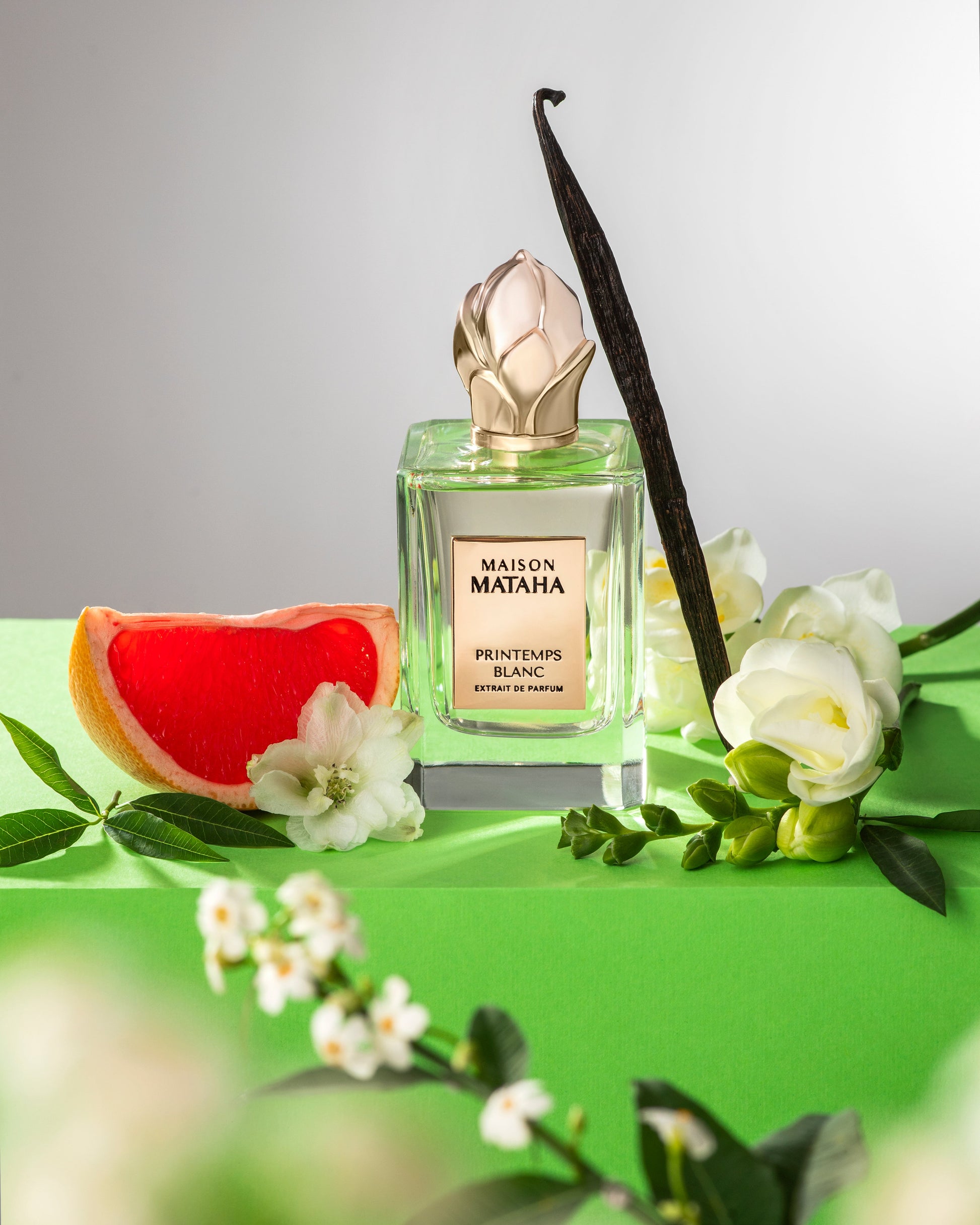 The Maison Mataha Printemps Blanc Extrait de Parfum bottle is shown with white flowers, jasmine sambac, vanilla bean, grapefruit slice, and green leaves on a bright green surface, reflecting its fresh floral scent.