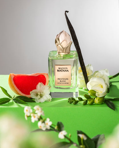 The Maison Mataha Printemps Blanc Extrait de Parfum bottle is shown with white flowers, jasmine sambac, vanilla bean, grapefruit slice, and green leaves on a bright green surface, reflecting its fresh floral scent.