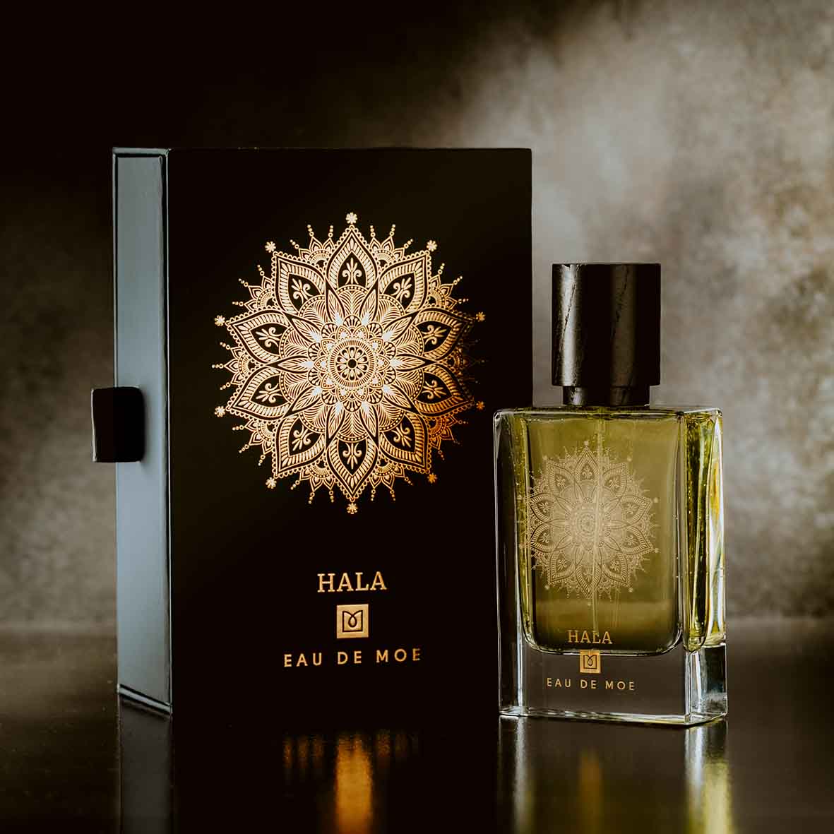 A HALA Eau de Moe perfume bottle, labeled by Eau De Moe, stands beside its matching black and gold box—both featuring an ornate gold mandala on a dark background—echoing the fruity floral scent inside.