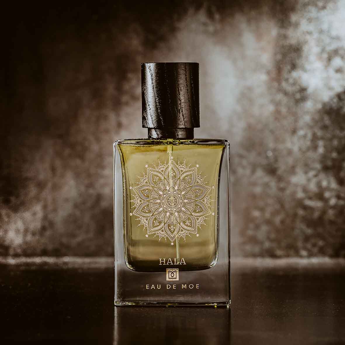 A glass perfume bottle labeled “HALA Eau de Moe” by Eau De Moe, featuring a black cap and mandala design, stands against a dark background, reflecting the fruity floral allure of this signature scent.