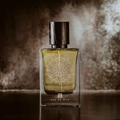 A glass perfume bottle labeled “HALA Eau de Moe” by Eau De Moe, featuring a black cap and mandala design, stands against a dark background, reflecting the fruity floral allure of this signature scent.