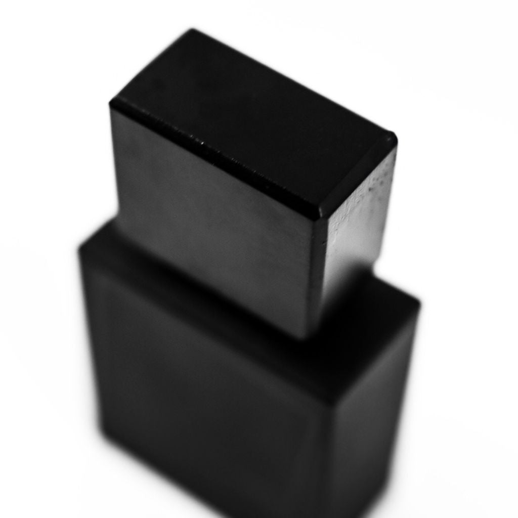 A close-up black and white photo of the rectangular, matte black UNUM LAVS EDP by Filippo Sorcinelli perfume bottle with a matching cap, set against a white background. The focus is on the cap, while the bottle appears slightly out of focus.