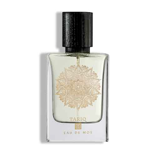 A clear glass bottle with a black cap, detailed gold mandala, and TARIQ Eau de Moe text. This Eau De Moe fragrance blends woody musk with vanilla, patchouli, and amber notes.
