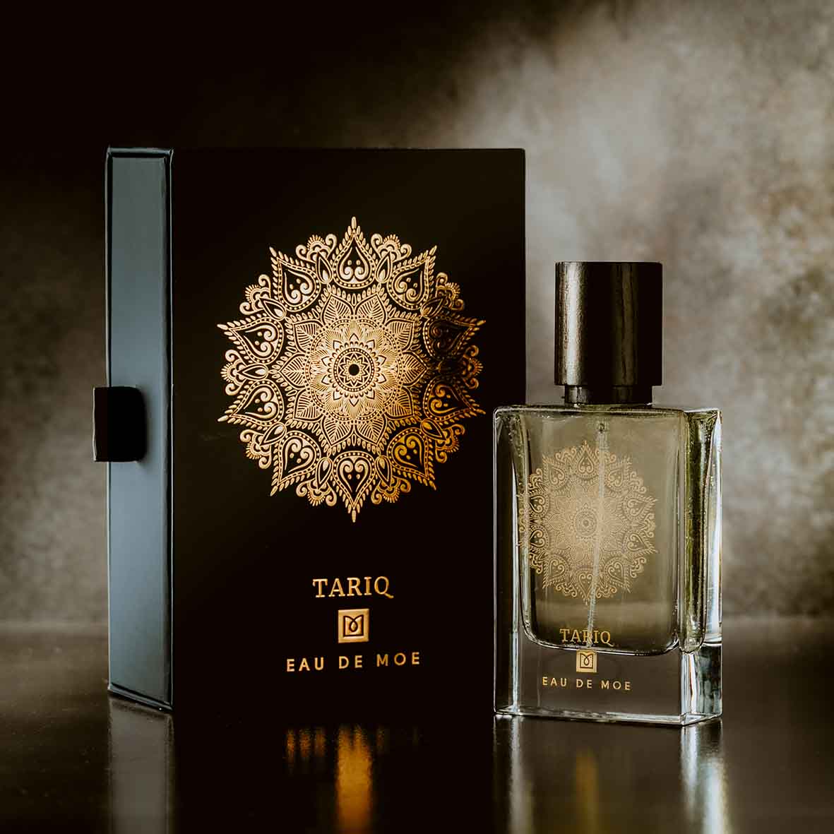 A stylish TARIQ Eau de Moe bottle by Eau De Moe stands beside its black and gold ornate box with a mandala design, set against an elegant background—highlighting its vanilla, patchouli, and amber notes.