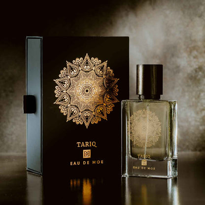 A stylish TARIQ Eau de Moe bottle by Eau De Moe stands beside its black and gold ornate box with a mandala design, set against an elegant background—highlighting its vanilla, patchouli, and amber notes.