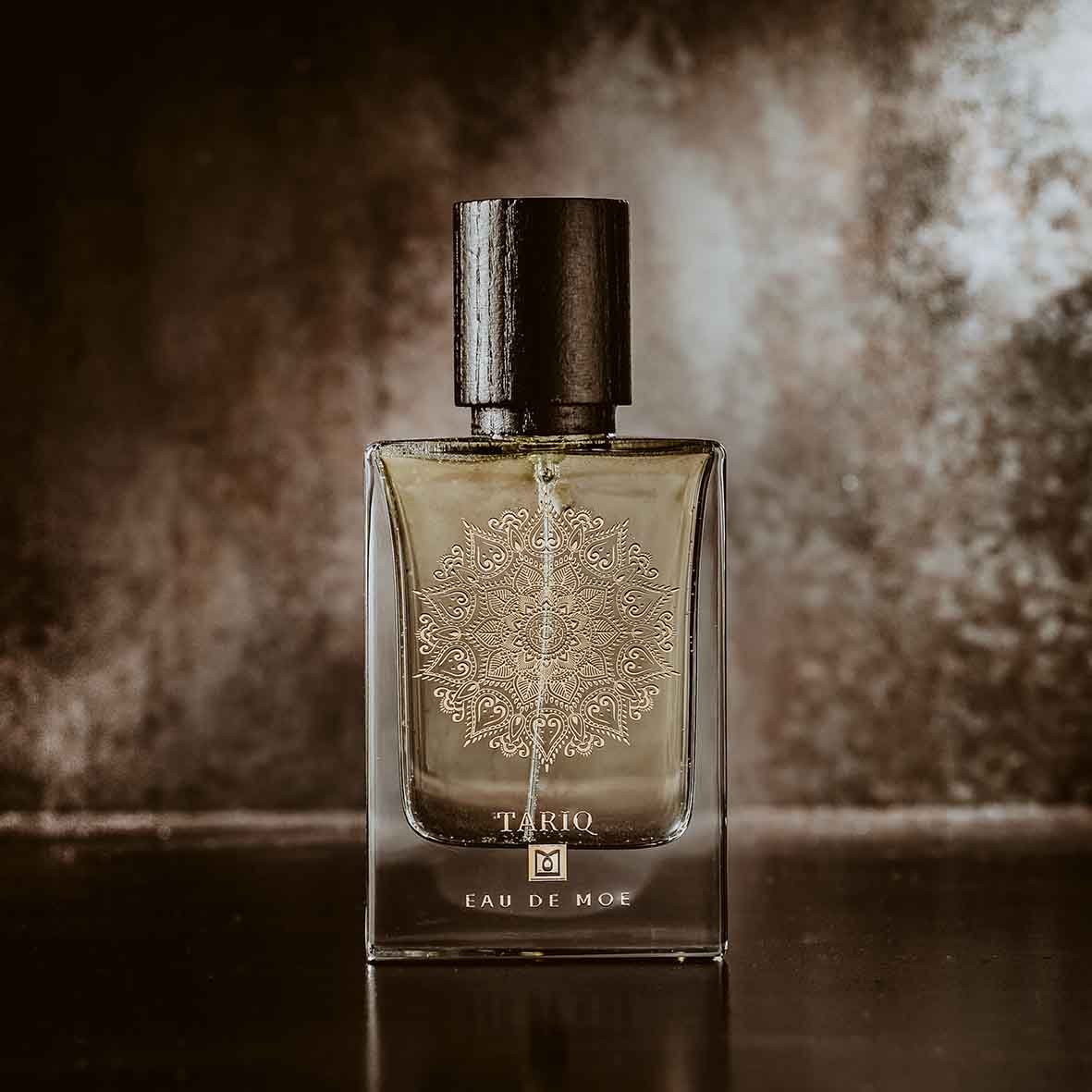 A glass TARIQ Eau de Moe perfume bottle by Eau De Moe, with a dark cap and mandala design, stands against a blurred background, evoking its woody musk scent of vanilla, patchouli, and amber.