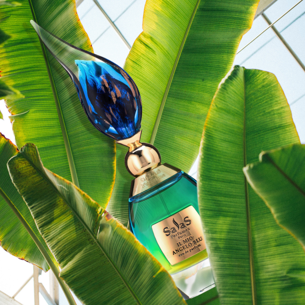 A blue and gold IL MIO ANGELO BLU EDP by Salas Art Perfumes, with its citrus-spice allure, stands among lush green tropical leaves as sunlight filters through a glass ceiling, evoking freshness and elegance.