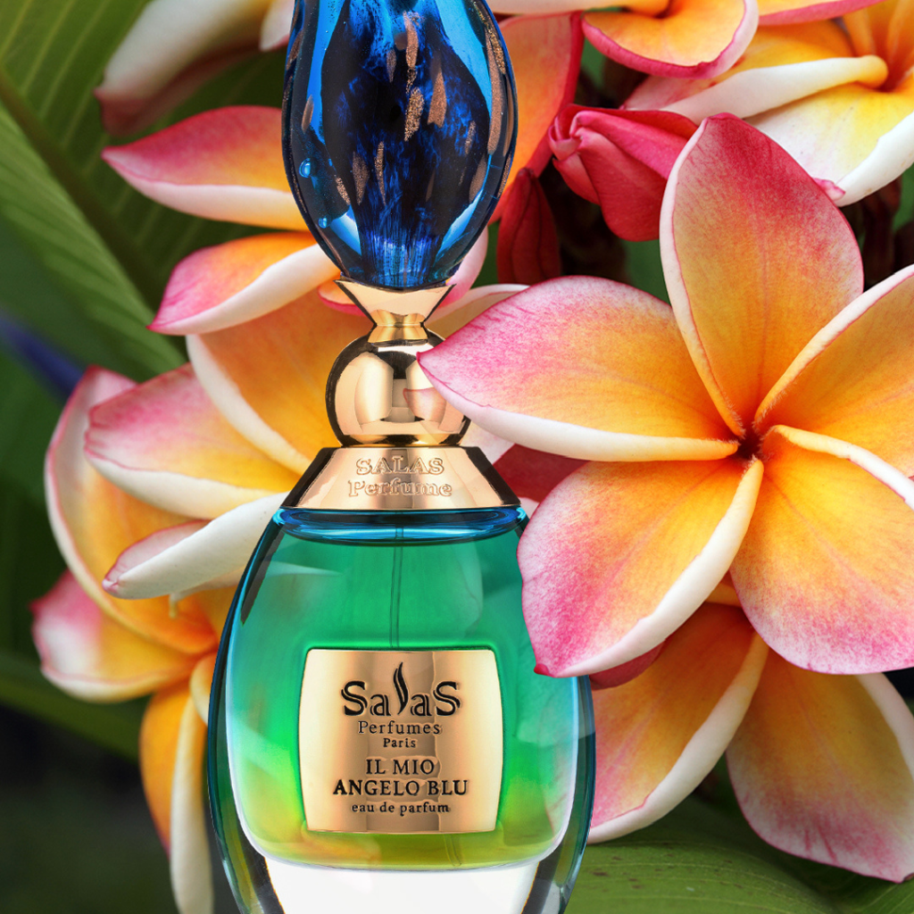 A green and blue Salas Art Perfumes IL MIO ANGELO BLU EDP bottle with citrus-spice notes sits before vibrant plumeria flowers in pink, yellow, and white, framed by lush green leaves.
