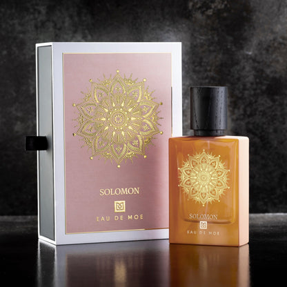 A bottle of SOLOMON Eau de Parfum by Eau De Moe, 80ML with a black cap, sits next to its decorative box—both adorned with a gold mandala on pink and gold backgrounds.