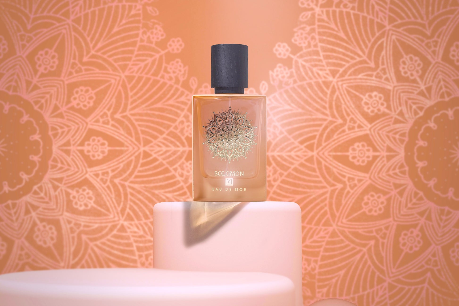 A bottle of SOLOMON Eau de Parfum by Eau De Moe sits on pale pink podiums, featuring a gold mandala design on the 80ML bottle and a matching peach-patterned background.