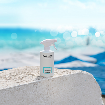 A white spray bottle labeled CASA MAR Sea Room Perfume by GIFTS sits on a white stone surface, with a bright blue ocean and sky blurred in the background.