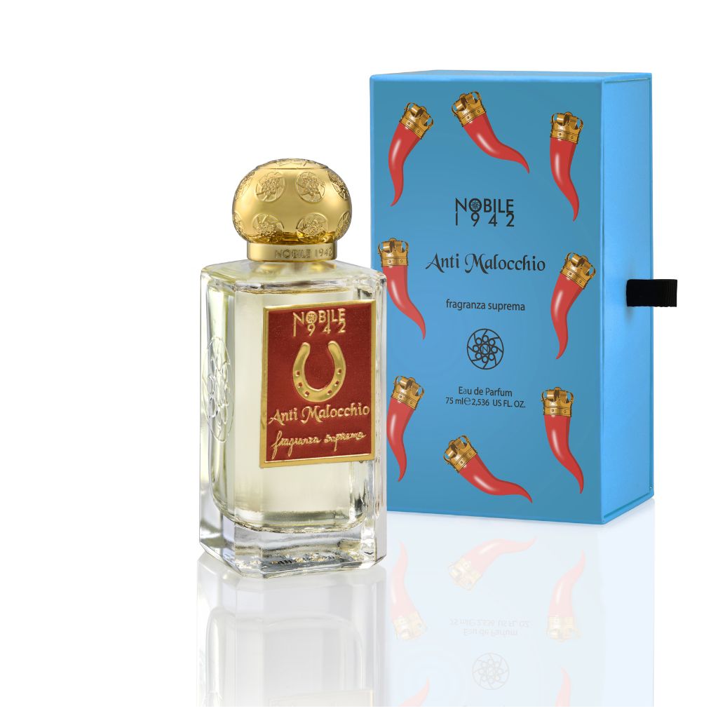 A clear glass ANTI MALOCCHIO Eau de Parfum bottle with a gold cap and red label by Nobile 1942 stands beside a blue box with gold details, coral charms, and amulets. The scent has citrus, aromatic, and woody notes.