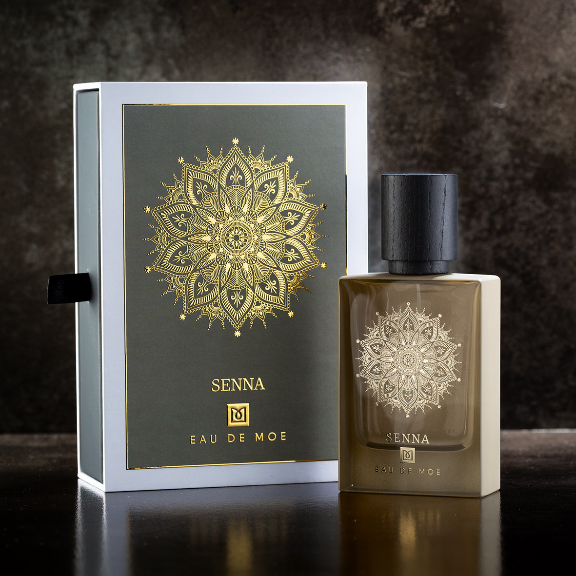 A bottle of SENNA Eau de Parfum by Eau De Moe sits next to its box, both adorned with a gold mandala on a dark backdrop and elegant black and gold text. The scent is sweet, powdery, and warmed by notes of Madagascar Vanilla.