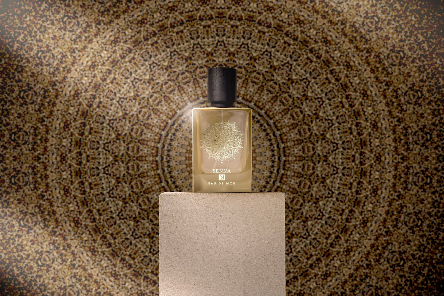 A SENNA Eau de Parfum bottle by Eau De Moe rests on a beige pedestal, framed by a golden mandala backdrop. The fragrance features enchanting Madagascar Vanilla for SENNA’s signature sweet, powdery scent.