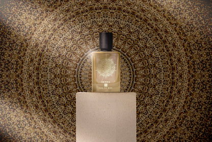 A SENNA Eau de Parfum bottle by Eau De Moe rests on a beige pedestal, framed by a golden mandala backdrop. The fragrance features enchanting Madagascar Vanilla for SENNA’s signature sweet, powdery scent.