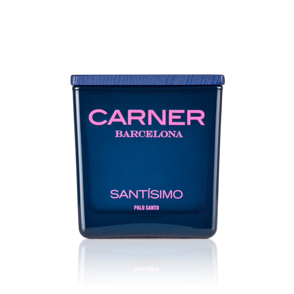 A blue SANTÍSIMO CANDLE by Carner features a dark blue lid. "CARNER" is in pink, and "SANTÍSIMO" in white and pink on the front, reflecting its guaiac wood-inspired scent.