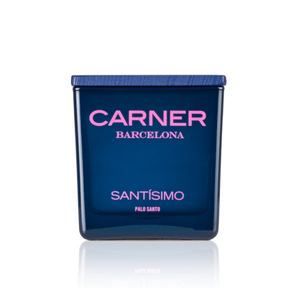 A blue SANTÍSIMO CANDLE by Carner features a dark blue lid. "CARNER" is in pink, and "SANTÍSIMO" in white and pink on the front, reflecting its guaiac wood-inspired scent.