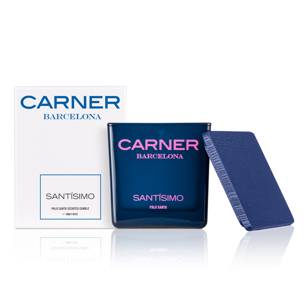 The SANTÍSIMO CANDLE by Carner Barcelona in dark blue sits beside its white box with blue and black text, both reflected on a surface and evoking musk and guaiac wood notes.
