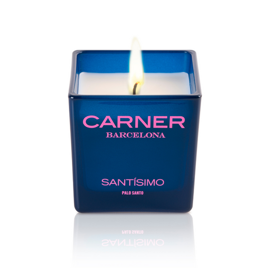 A lit SANTÍSIMO CANDLE by Carner, in a blue holder labeled Carner Barcelona Santísimo, stands on a white reflective surface; the scent features notes of guaiac wood.