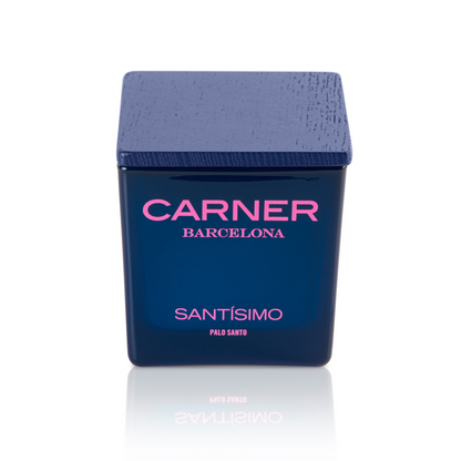 A dark blue SANTÍSIMO CANDLE by Carner Barcelona, with a textured square lid and pink and white label, highlights musk and guaiac wood notes, displayed against a white background.