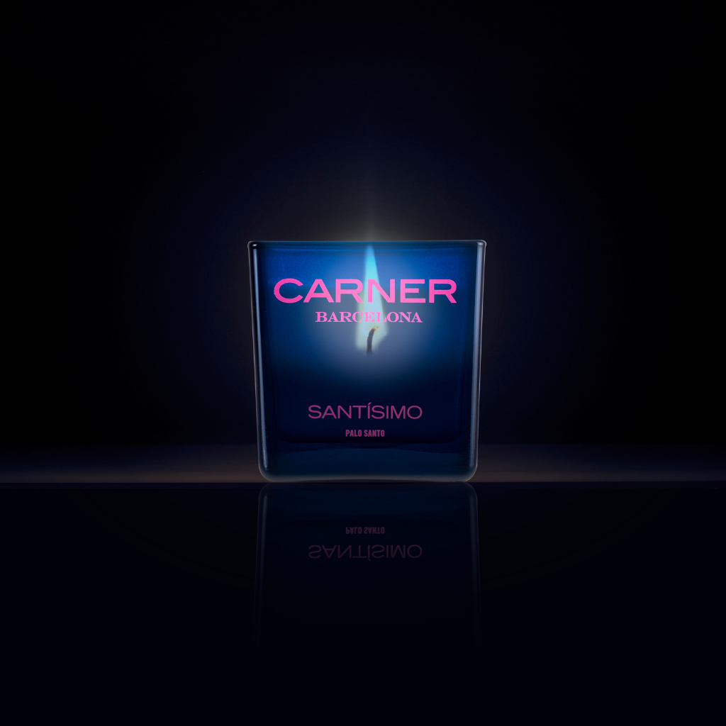 A dark blue SANTÍSIMO CANDLE by Carner burns brightly against a black backdrop with a soft blue glow and mirrored reflection, filling the air with notes of musk and guaiac wood.