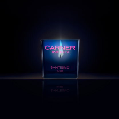 A dark blue SANTÍSIMO CANDLE by Carner burns brightly against a black backdrop with a soft blue glow and mirrored reflection, filling the air with notes of musk and guaiac wood.