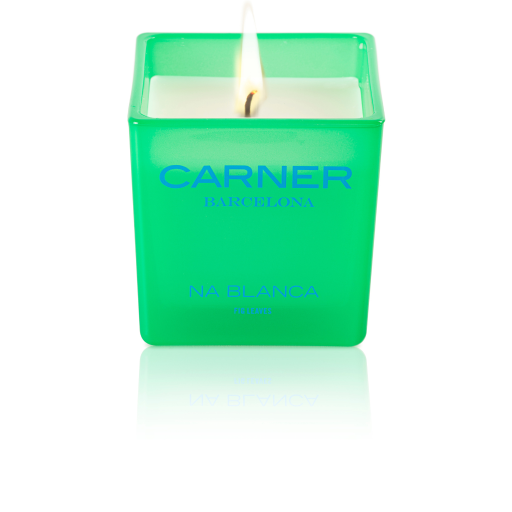 A lit NA BLANCA CANDLE by GIFTS in a green square glass holder evokes the essence of fig trees on Formentera, with its white wax glowing and a subtle reflection beneath.