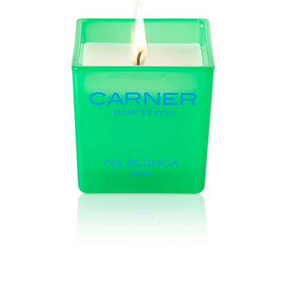 A lit NA BLANCA CANDLE by GIFTS in a green square glass holder evokes the essence of fig trees on Formentera, with its white wax glowing and a subtle reflection beneath.