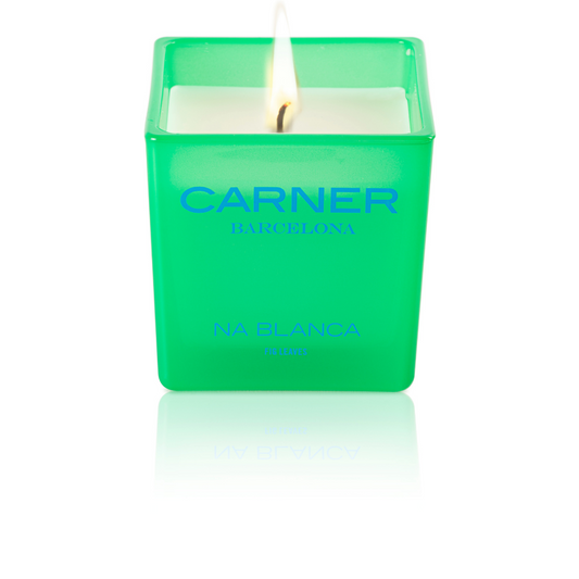 A lit NA BLANCA CANDLE by GIFTS in a green square glass holder evokes the essence of fig trees on Formentera, with its white wax glowing and a subtle reflection beneath.
