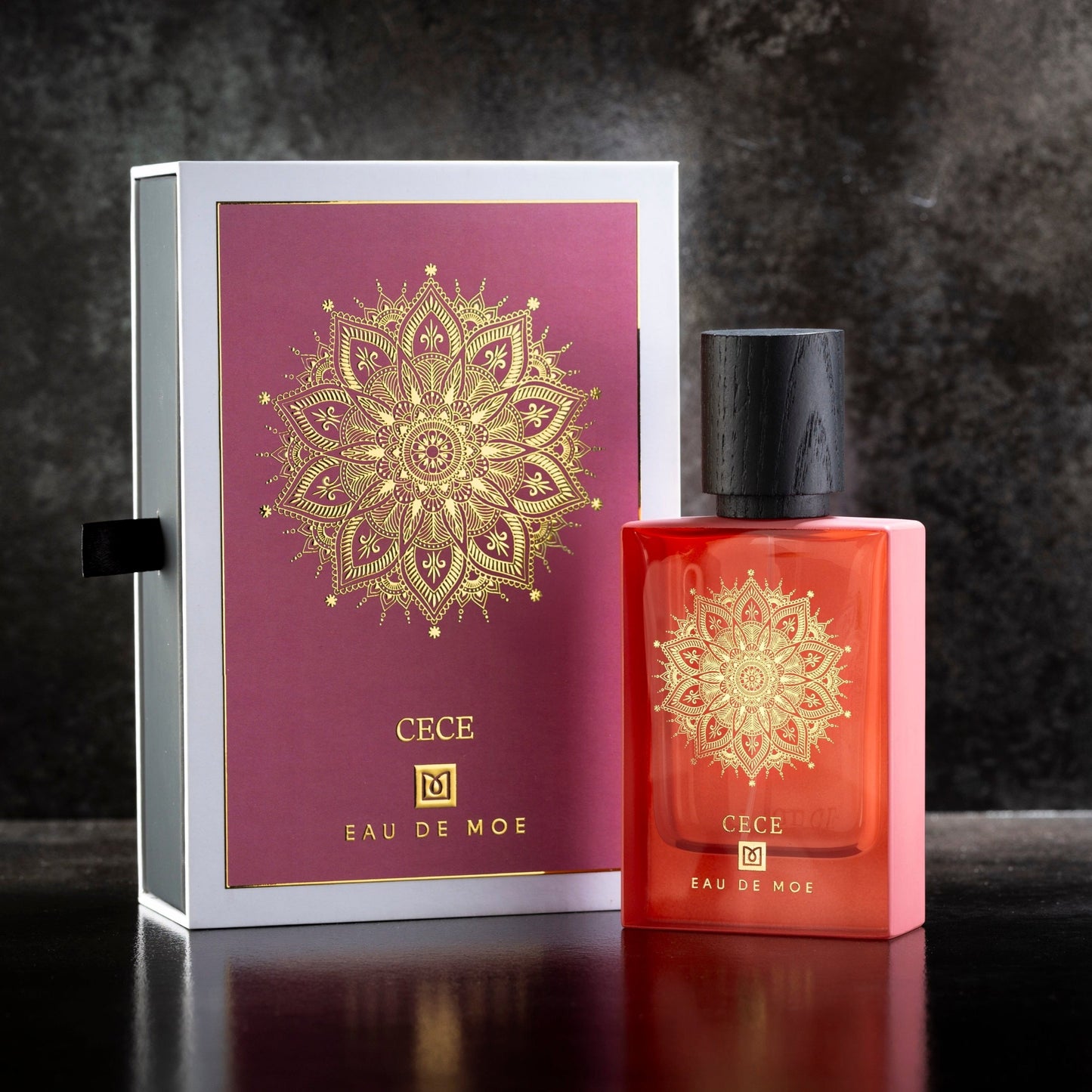 A red CECE Eau de Parfum bottle by Eau De Moe, featuring a gold mandala design and black cap, stands beside its matching box on a dark surface.