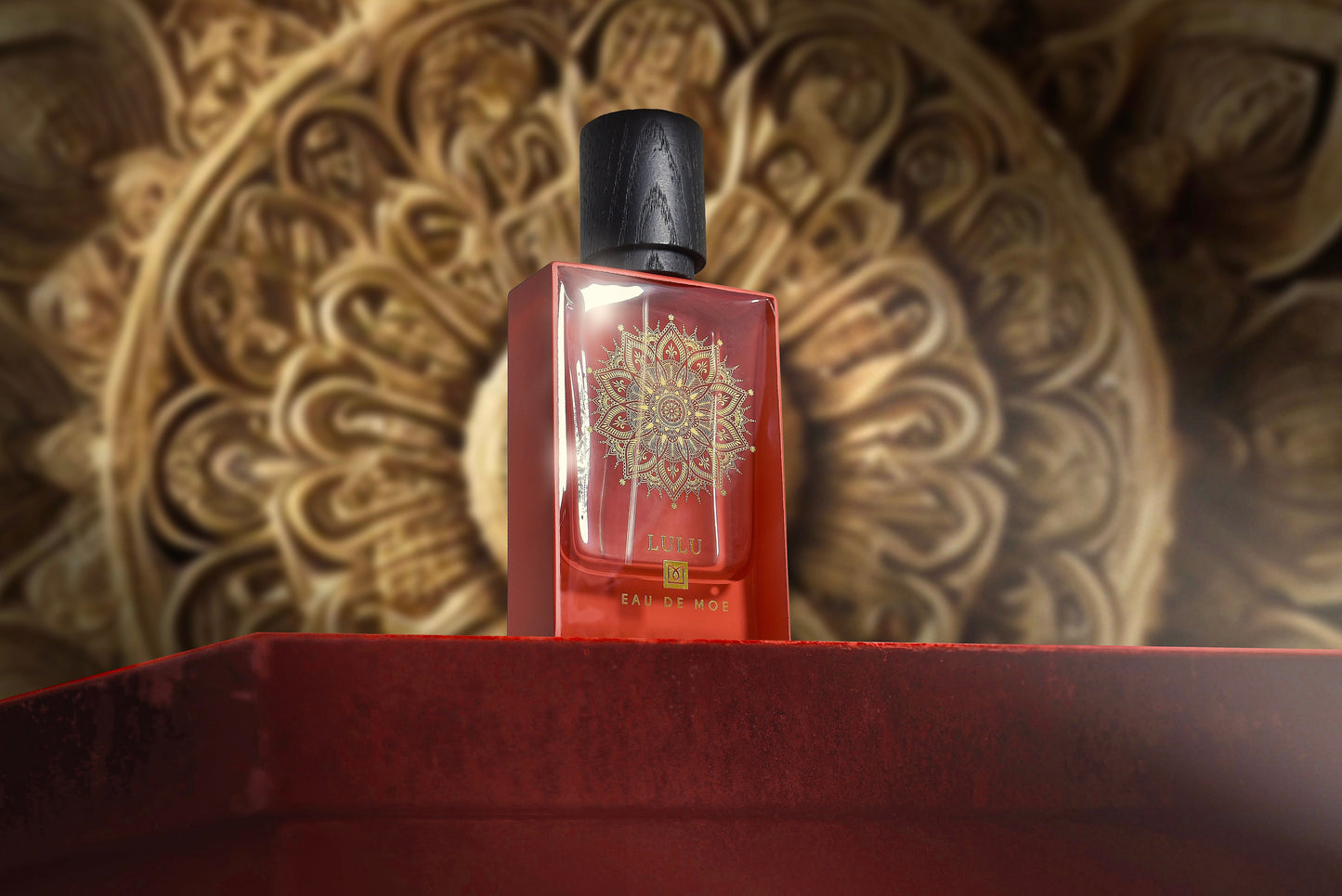 A red LULU Eau de Parfum bottle by Eau De Moe, featuring a gold mandala and black cap, sits on a red surface with an ornate blurred background—a striking 80ML women’s fragrance.