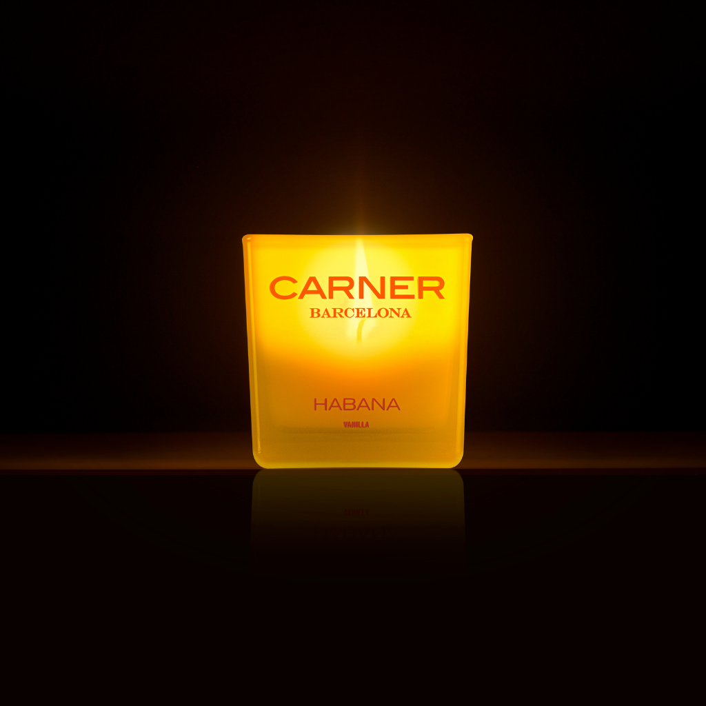 A glowing HABANA CANDLE by GIFTS, scented and set in a frosted glass holder with Carner Barcelona branding, shines warmly against a dark backdrop.