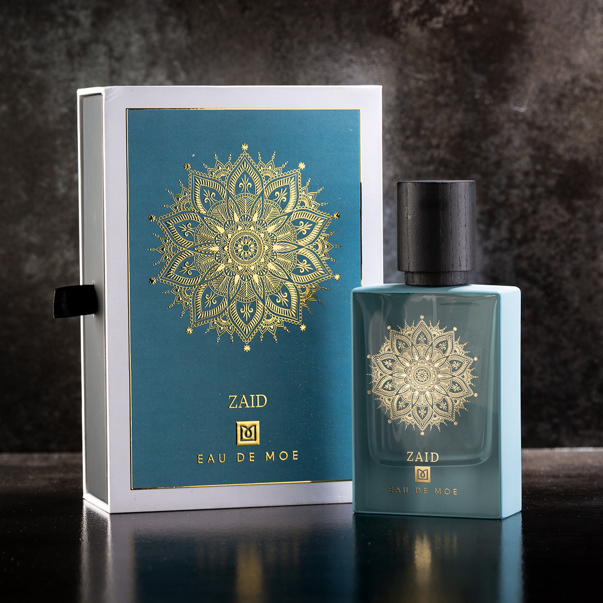 A blue ZAID Eau de Parfum 80ML bottle by Eau De Moe stands before its matching box, both featuring a gold mandala on teal, set against a dark backdrop. Experience the allure of ZAID Eau de Parfum.