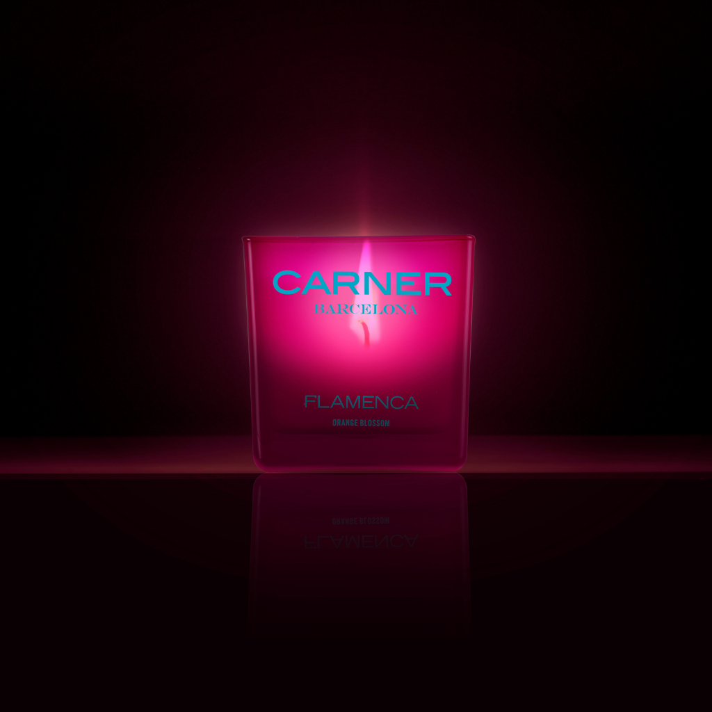 The FLAMENCA CANDLE - Carner Barcelona by GIFTS glows pink with notes of orange blossom and bergamot, standing against a dark background and casting a soft light and reflection below.