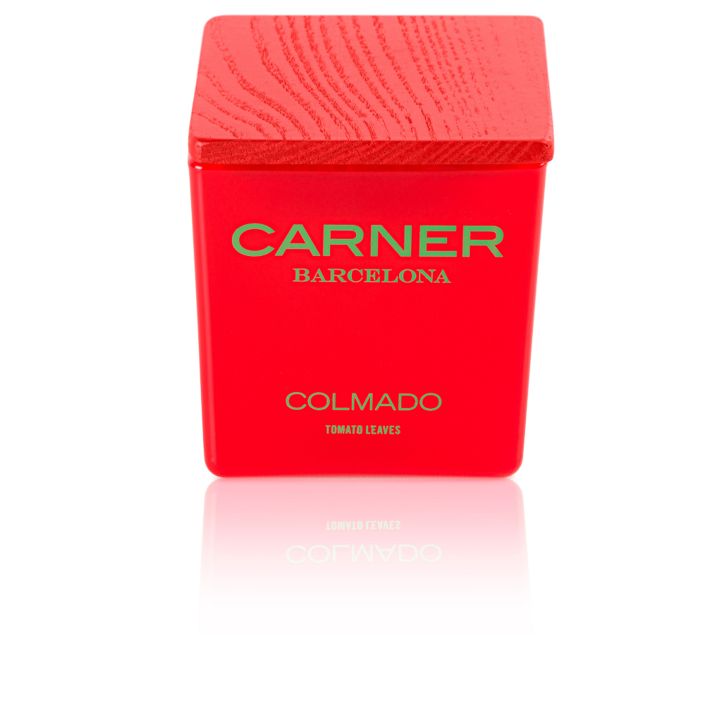 A red square “COLMADO CANDLE - Carner Barcelona” by GIFTS, featuring a wooden-textured lid and green text, is inspired by colmados and shown brightly lit against a white background.