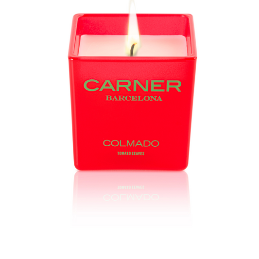 A lit COLMADO CANDLE by GIFTS (Carner Barcelona) glows above white wax in a square container labeled TOMATO LEAVES.