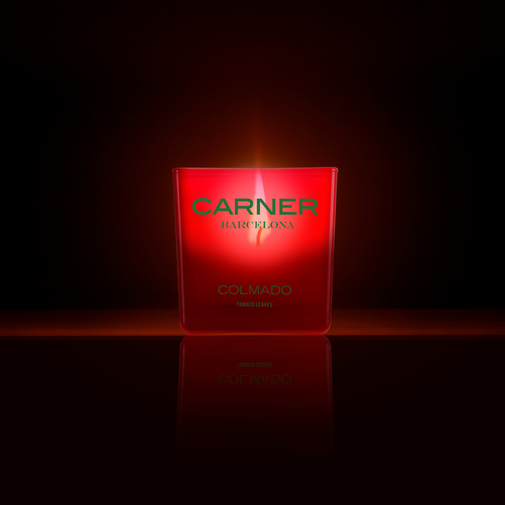 A glowing red COLMADO CANDLE by Carner Barcelona (GIFTS) sits against a dark background, its flame softly illuminating the container and casting a gentle reflection below.